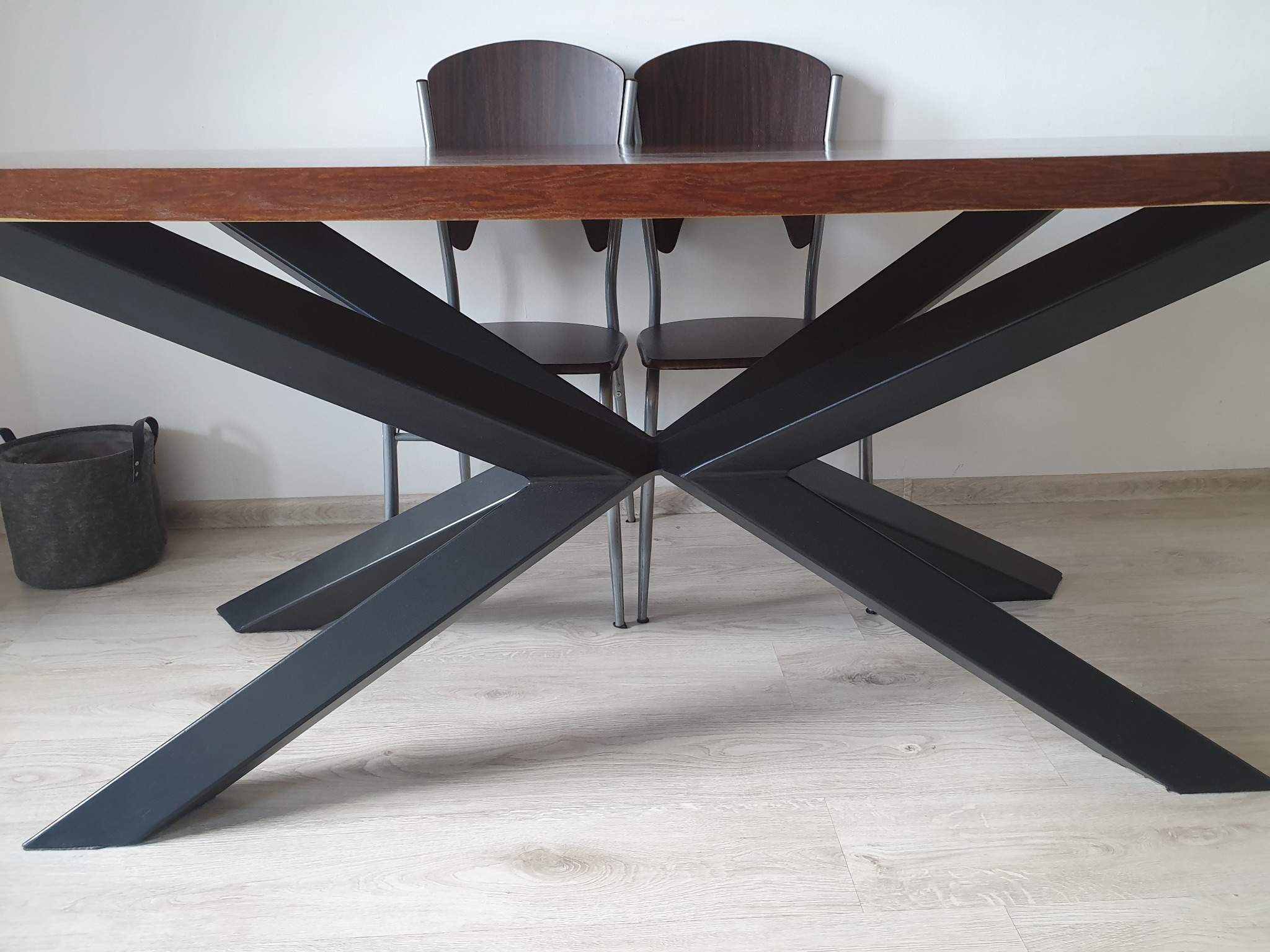 Stół z blatem z ciemnego drewna i czarną, metalową podstawą w kształcie litery X, widziane z dołu, z dwoma krzesłami w tle i koszem z lewej strony.