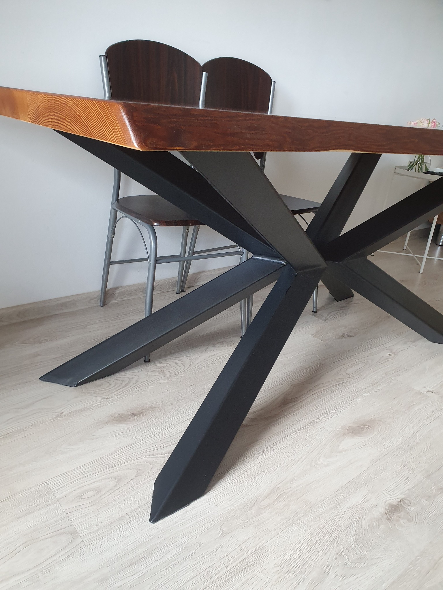 Stół z blatem z jasnego drewna i czarną, metalową podstawą w kształcie gwiazdy, widoczne krzesła w tle.