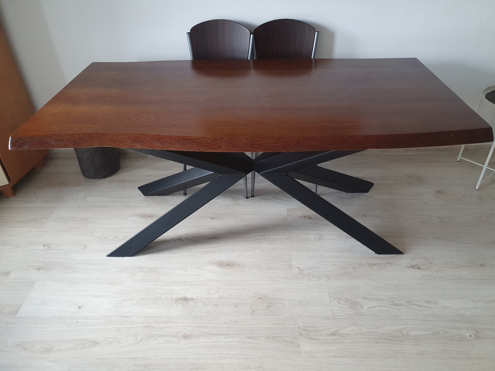 Stół z blatem z ciemnego drewna o nieregularnych krawędziach, wsparty na czarnej, geometrycznej podstawie, ustawiony na jasnej podłodze, z dwoma krzesłami w tle.