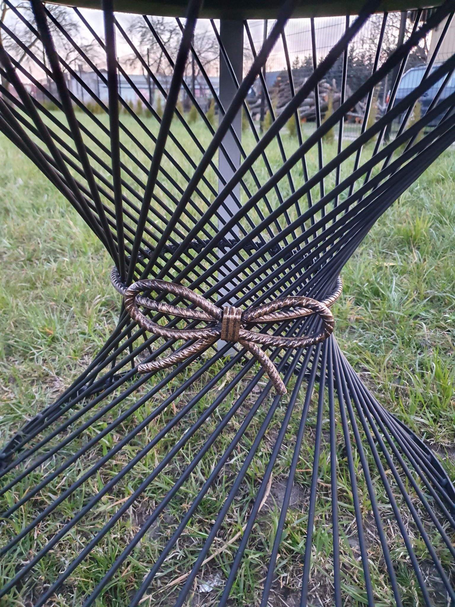 Metalowa konstrukcja stołu ogrodowego z ozdobnym wiązaniem w kształcie kokardy, widoczna na tle trawnika.