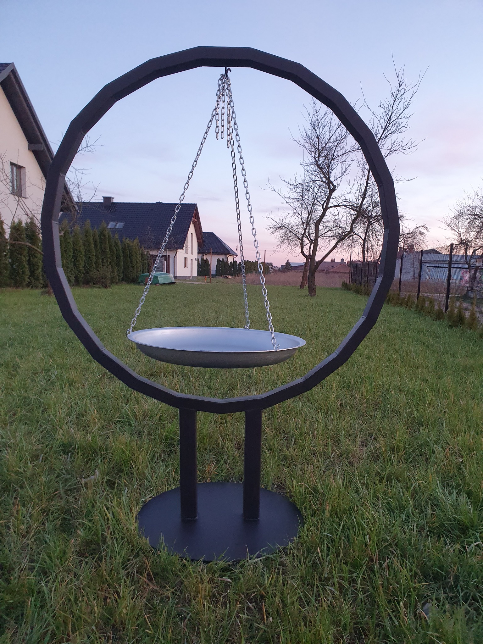 Metalowa konstrukcja w kształcie okręgu z podwieszaną misą na łańcuchach, stojąca na trawniku przed domem jednorodzinnym o zmierzchu.