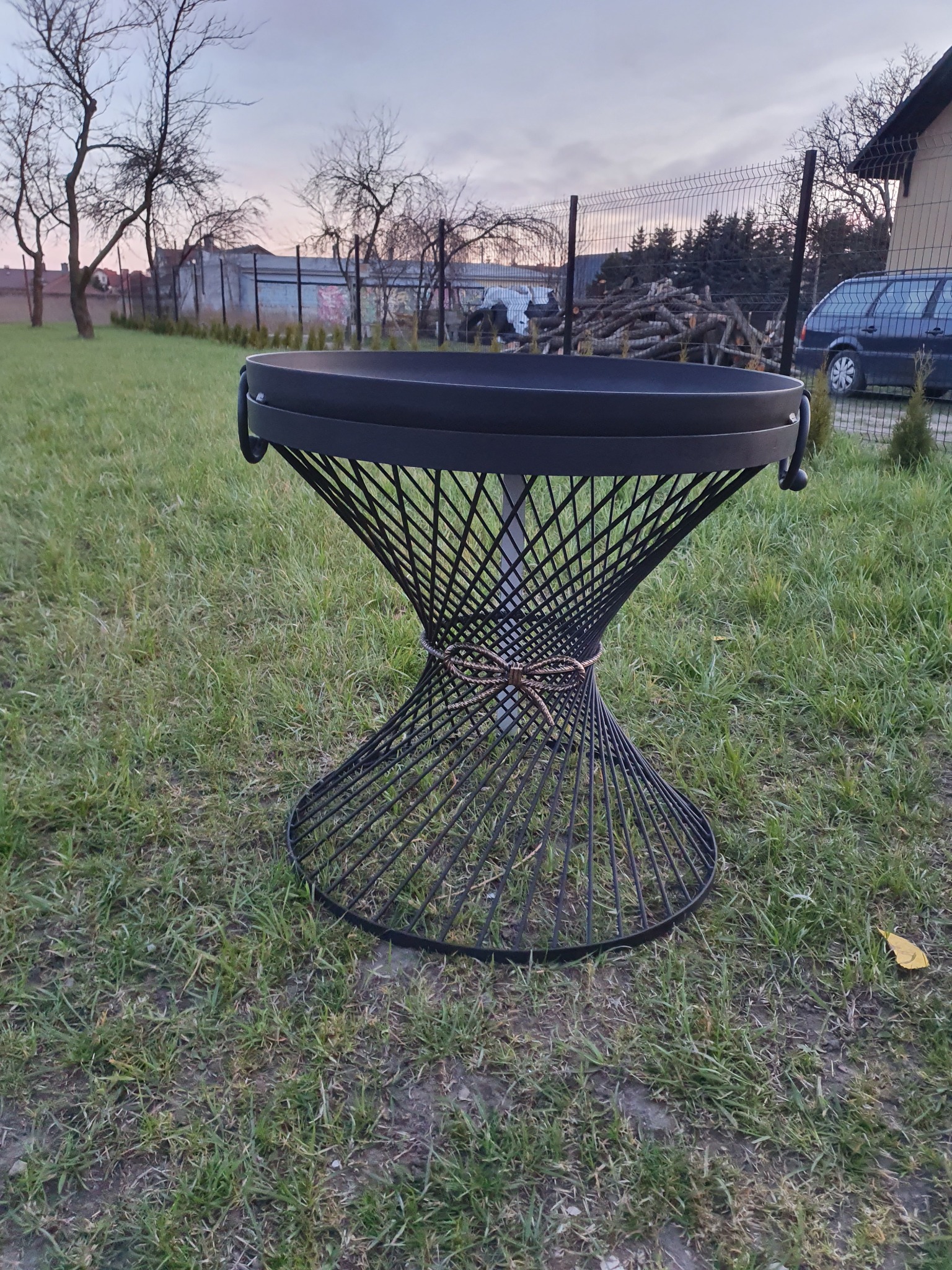 Czarny, metalowy kosz na ognisko o ażurowej, zwężającej się ku dołowi konstrukcji, ustawiony na trawniku w ogrodzie, z widocznymi drzewami i ogrodzeniem w tle.