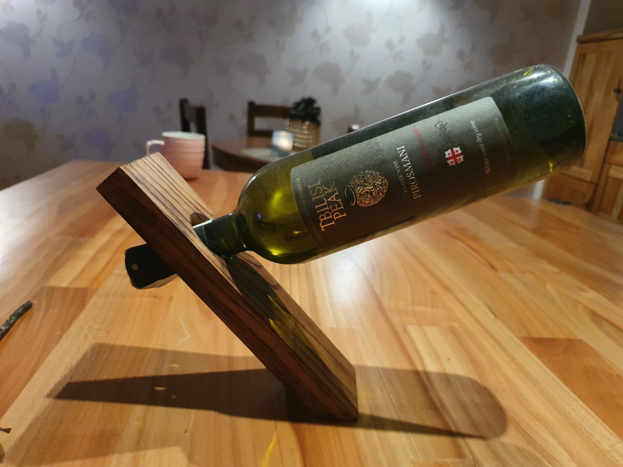Butelka wina Tbilisi Peak Pirosmani leżąca pod kątem na drewnianym stojaku na stole z drewna. W tle widoczny fragment ściany z kwiatowym wzorem, różowy kubek i fragment drewnianego krzesła.