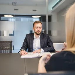 Mężczyzna w garniturze siedzi przy stole w biurze, rozmawiając z kobietą trzymającą biały kubek; na stole otwarty dokument.