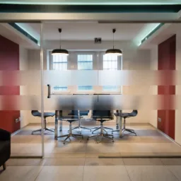 Szklana ściana biurowa z poziomymi pasami matowego szkła oddzielająca salę konferencyjną z metalowymi krzesłami i stołem, widoczny fragment sofy w recepcji.