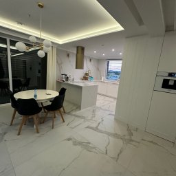 Comfort Lux - Nowoczesna kuchnia z marmurową podłogą i ścianą, okrągły stół z czarnymi krzesłami, złoty okap nad wyspą kuchenną, oświetlenie LED w suficie.