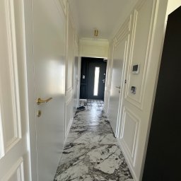 Comfort Lux - Korytarz z białymi drzwiami panelowymi, marmurową podłogą i czarnymi drzwiami wejściowymi w głębi.