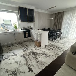 Comfort Lux - Nowoczesna kuchnia z marmurową podłogą, białymi szafkami i czarnym stołem jadalnym, doświetlona naturalnym światłem wpadającym przez okno.