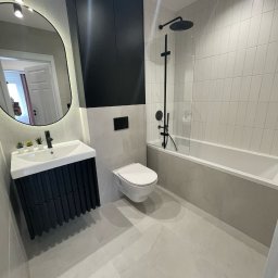 Comfort Lux - Nowoczesna łazienka z szarymi płytkami, czarną armaturą i szafką umywalkową z lamelami, owalne lustro z podświetleniem LED, minimalistyczny design.