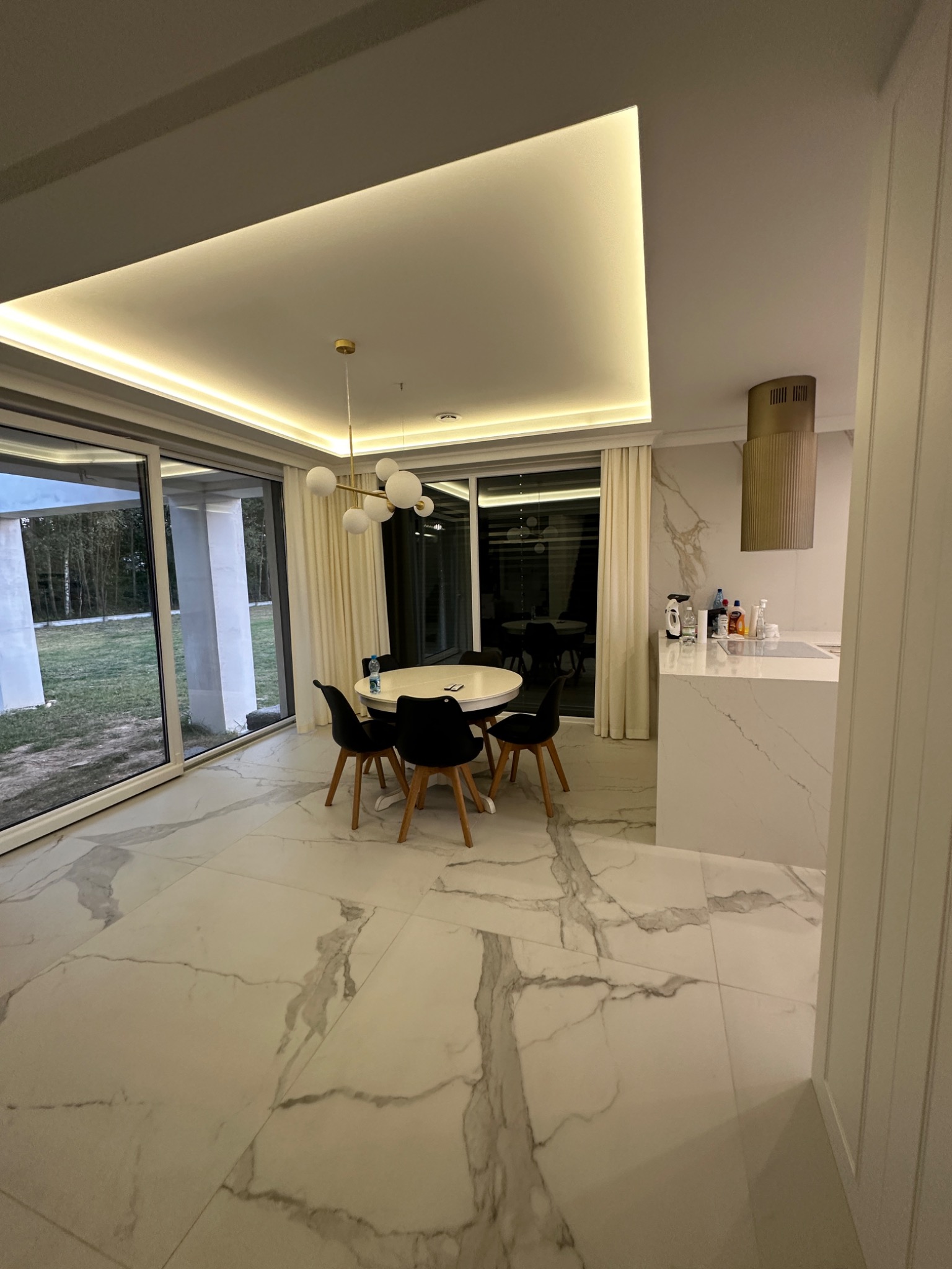 Nowoczesna jadalnia z marmurową podłogą i blatem kuchennym, oświetlenie LED w suficie, okno panoramiczne z widokiem na zieleń.