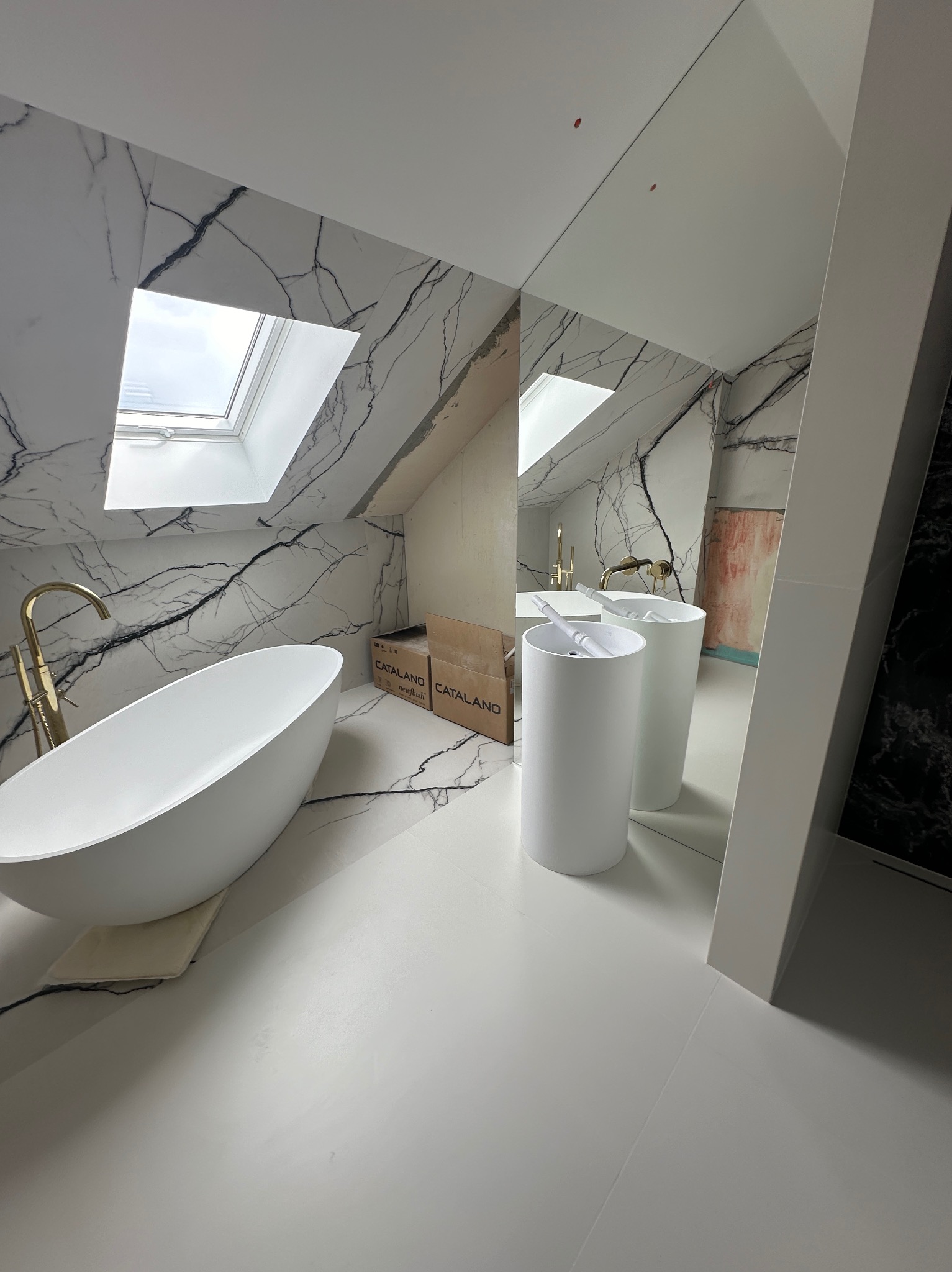 Nowoczesna łazienka na poddaszu z wanną wolnostojącą, umywalkami kolumnowymi i marmurowymi płytkami na ścianach, widoczne kartony po ceramice.