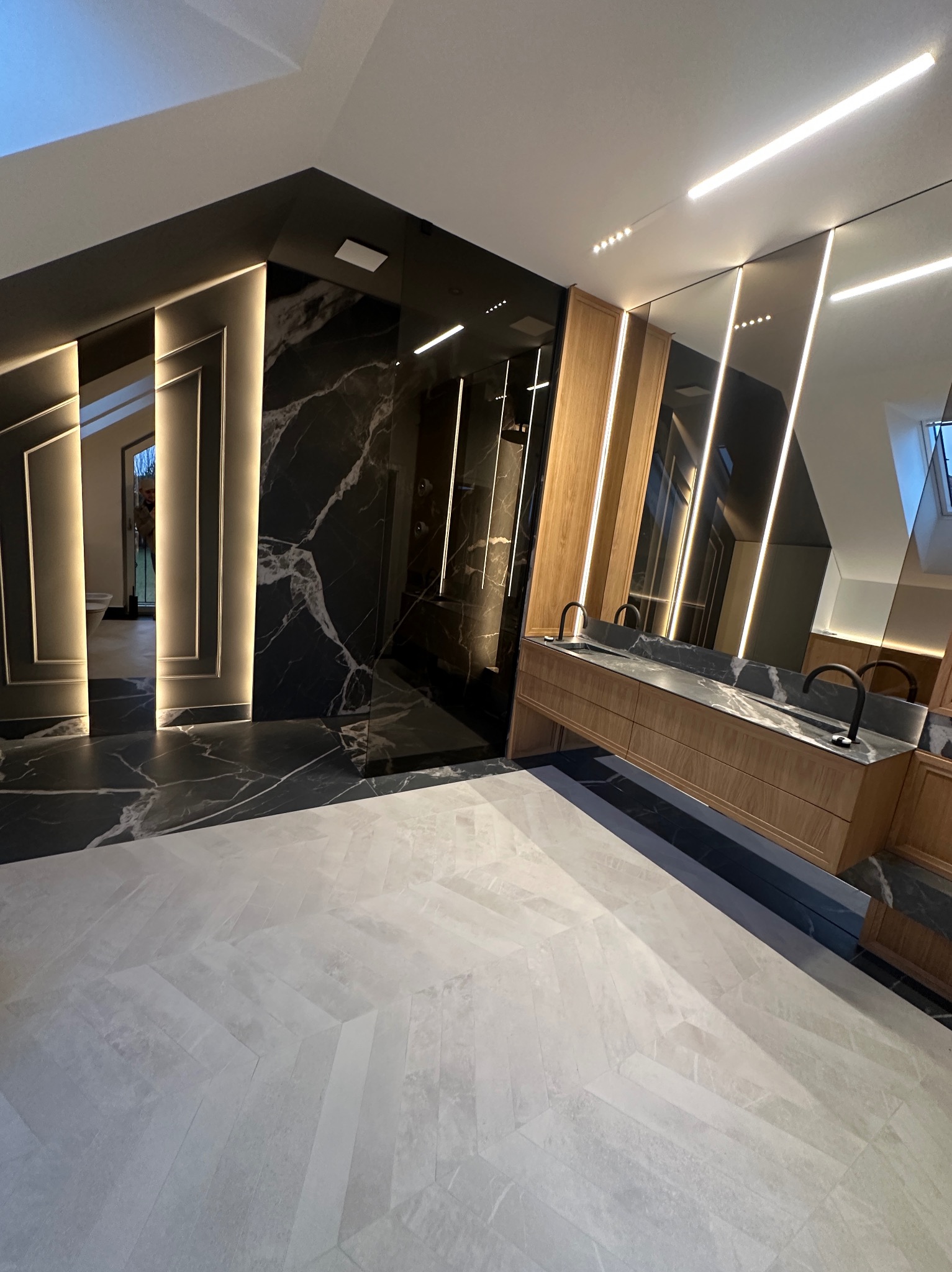 Nowoczesna łazienka z czarnymi marmurowymi ścianami i podłogą, drewnianą szafką pod umywalkę i dużym lustrem z oświetleniem LED, jasne płytki podłogowe ułożone we wzór jodełki.