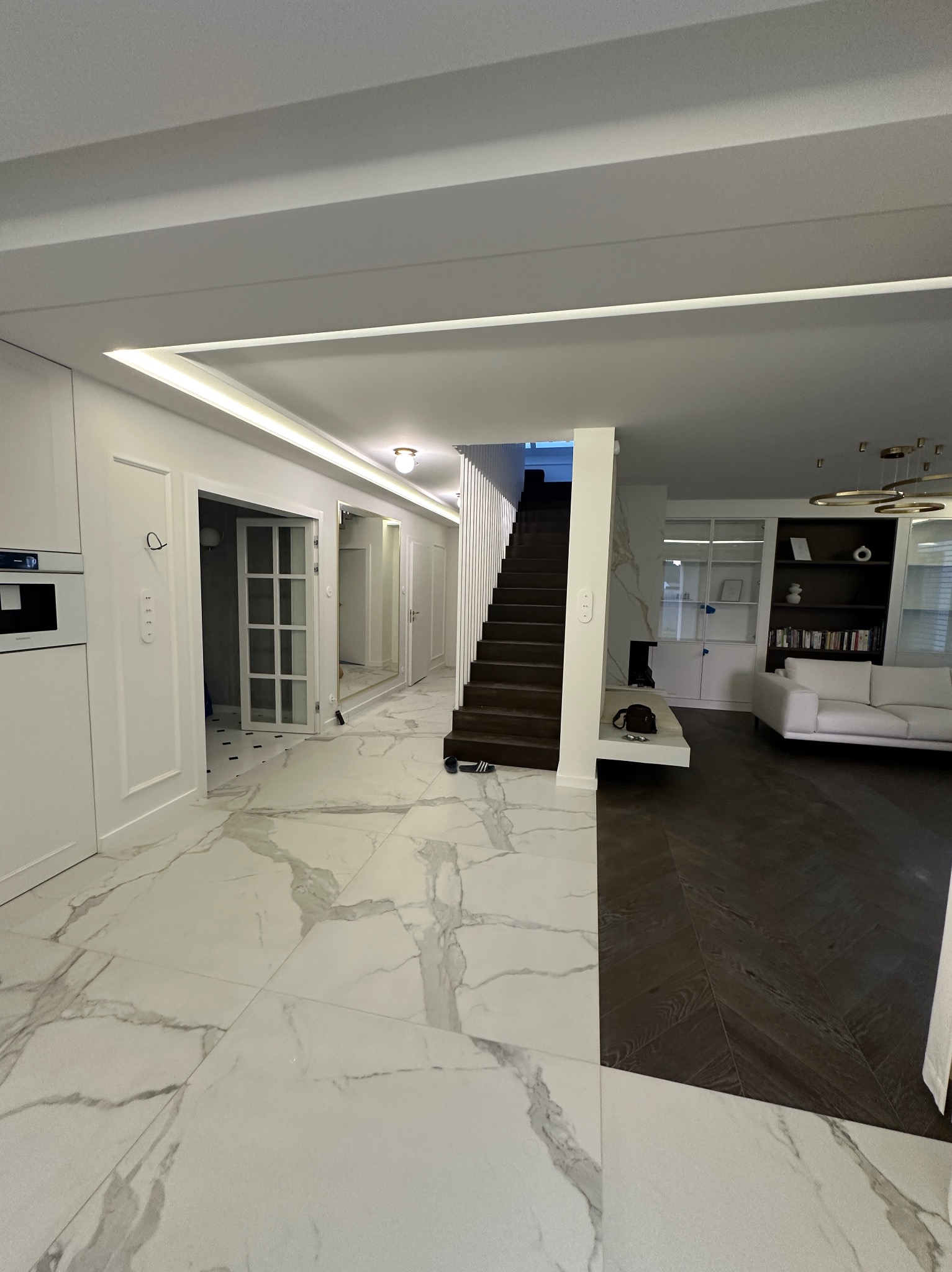 Nowoczesny hol z białymi marmurowymi płytkami podłogowymi kontrastującymi z ciemną drewnianą podłogą ułożoną w jodełkę, białe ściany z dekoracyjnymi listwami, schody z ciemnego drewna...