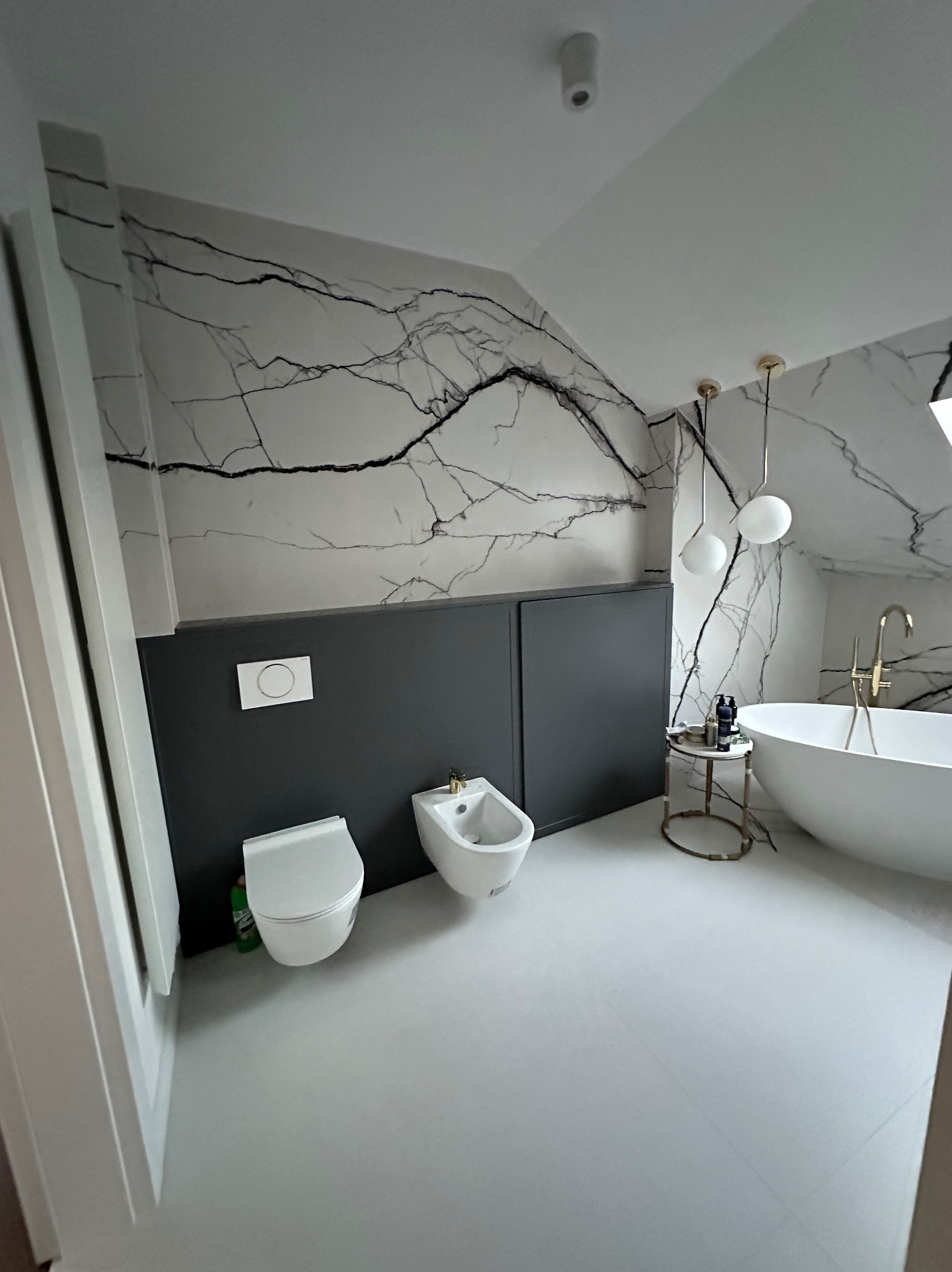 Nowoczesna łazienka z białą ceramiką, wanną wolnostojącą z złotą armaturą, ścianami wykończonymi płytami imitującymi marmur z czarnymi żyłami i szarą lamperią.