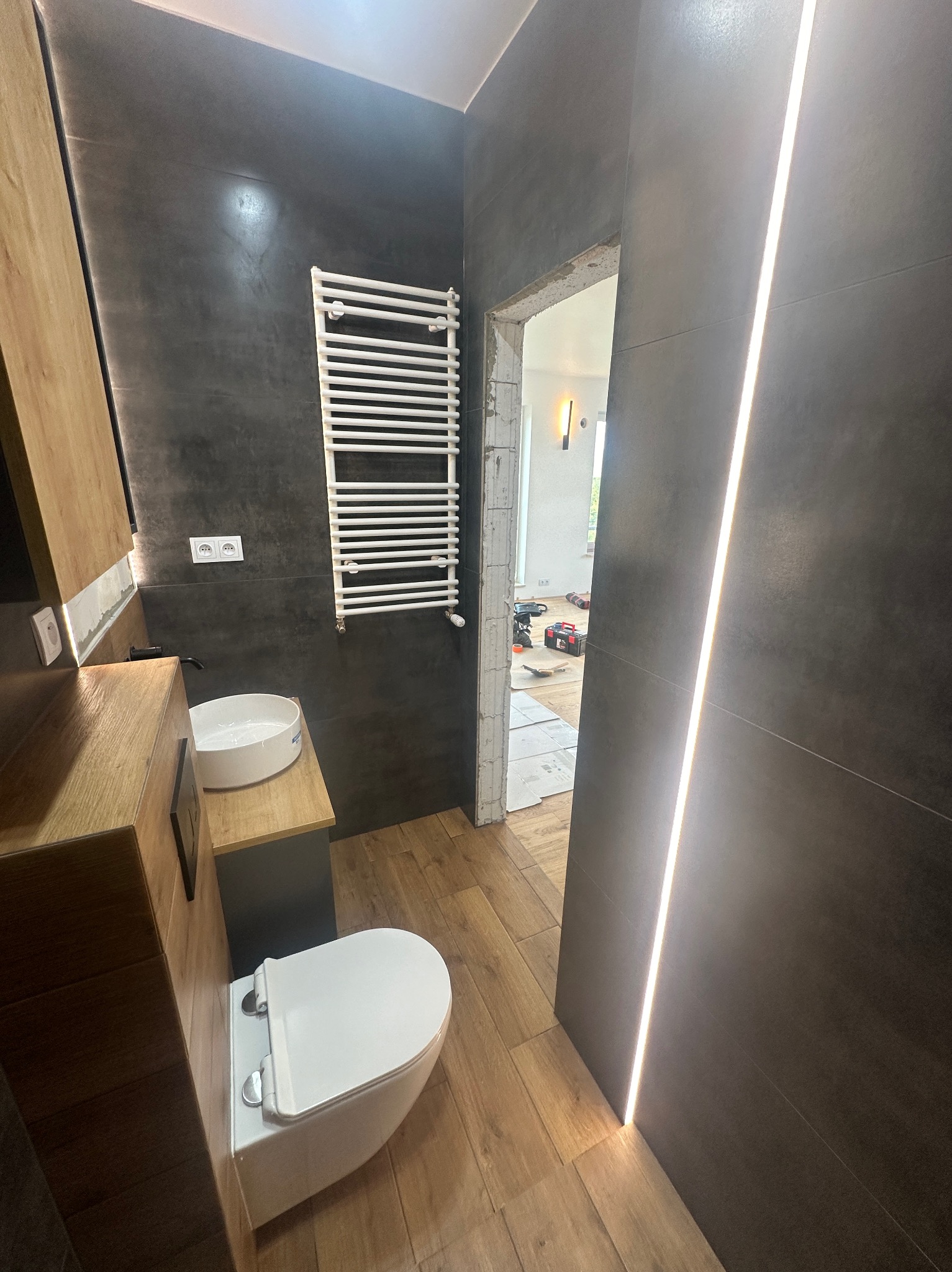 Ciemnoszara łazienka z drewnianymi akcentami, podświetleniem LED wzdłuż ściany, białą umywalką i toaletą, oraz grzejnikiem drabinkowym; widoczny fragment remontowanego pomieszczenia obok.