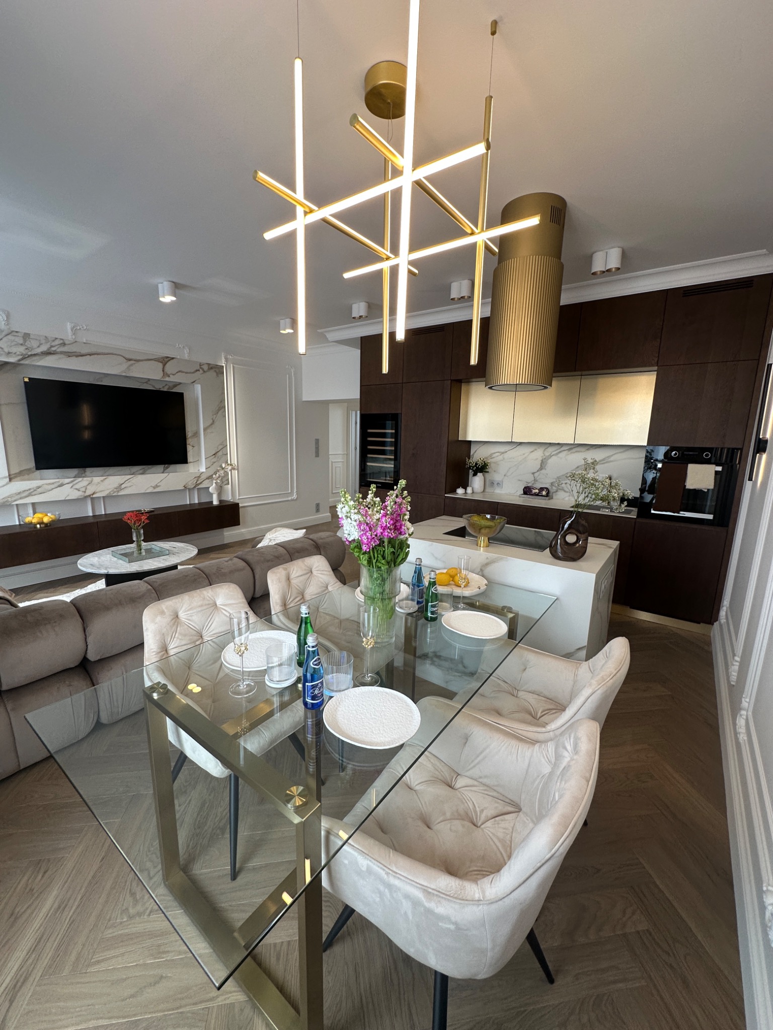 Nowoczesne wnętrze salonu z aneksem kuchennym, szklany stół z beżowymi krzesłami, designerska lampa wisząca, parkiet ułożony w jodełkę, ściana z marmurowym wykończeniem.
