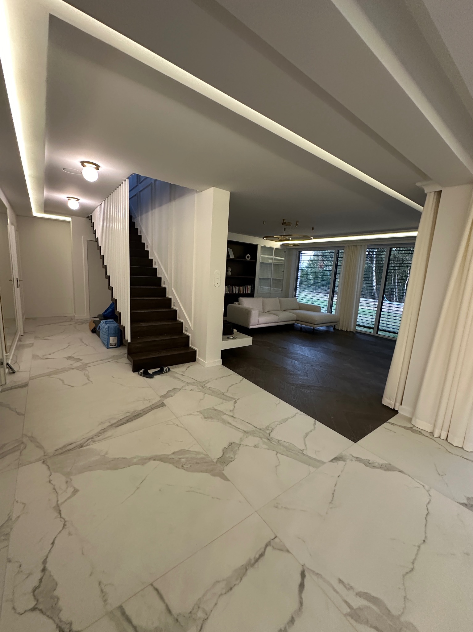 Przestronne wnętrze domu z marmurową podłogą, drewnianymi schodami i salonem z białymi meblami, oświetlone listwami LED w suficie.