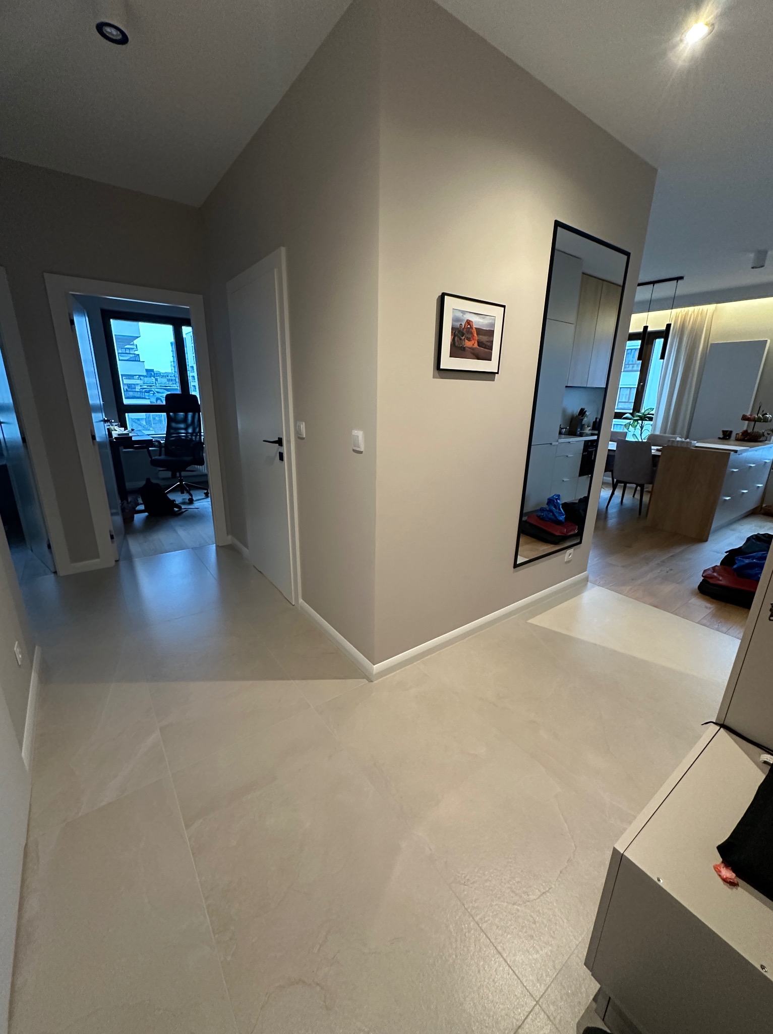 Beżowe płytki podłogowe w korytarzu mieszkania z widokiem na biuro i kuchnię, ściany pomalowane na jasny odcień szarości, lustro w czarnej ramie.