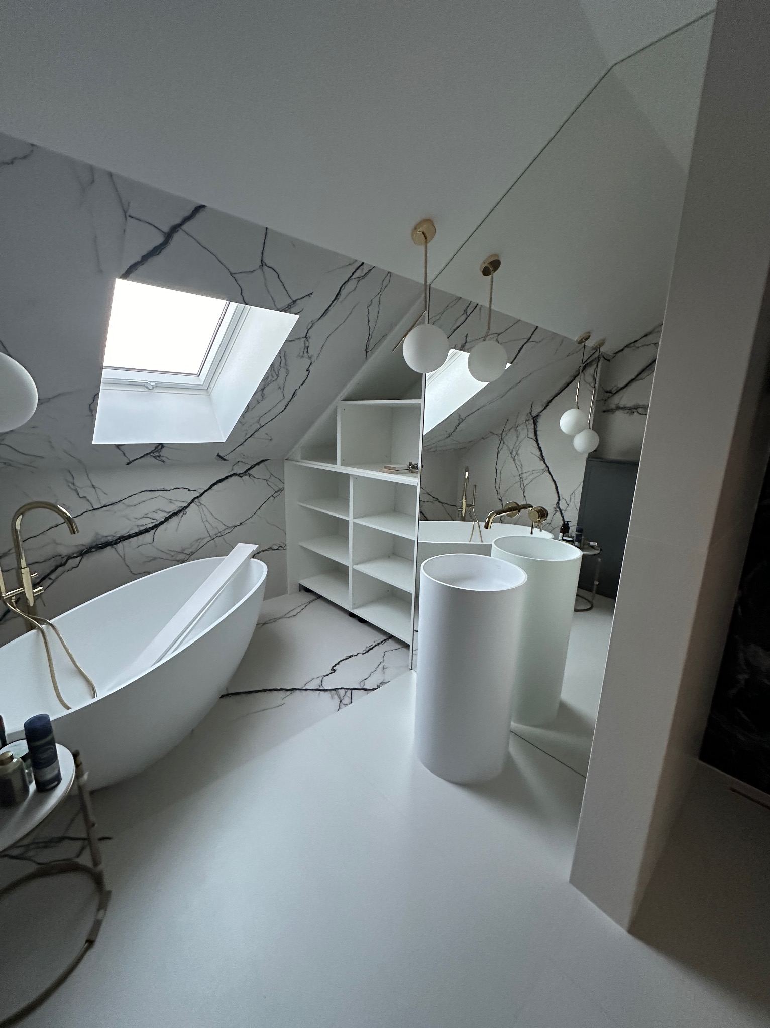 Nowoczesna łazienka na poddaszu z białą wolnostojącą wanną, umywalkami na postumentach, złotą armaturą i marmurowymi płytkami na ścianach pod skosem z oknem dachowym.