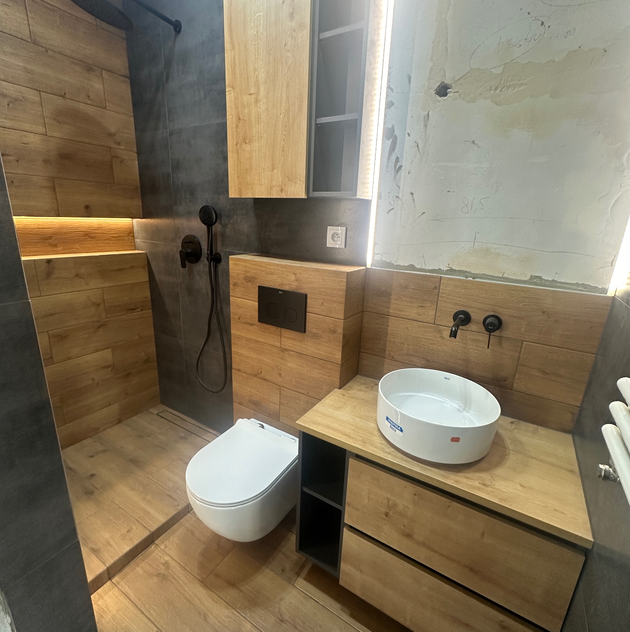 Nowoczesna łazienka z drewnopodobnymi płytkami, czarną armaturą i podświetleniem LED, ukazująca umywalkę nablatową i toaletę podwieszaną, z widocznymi niedoskonałościami ściany w tle.