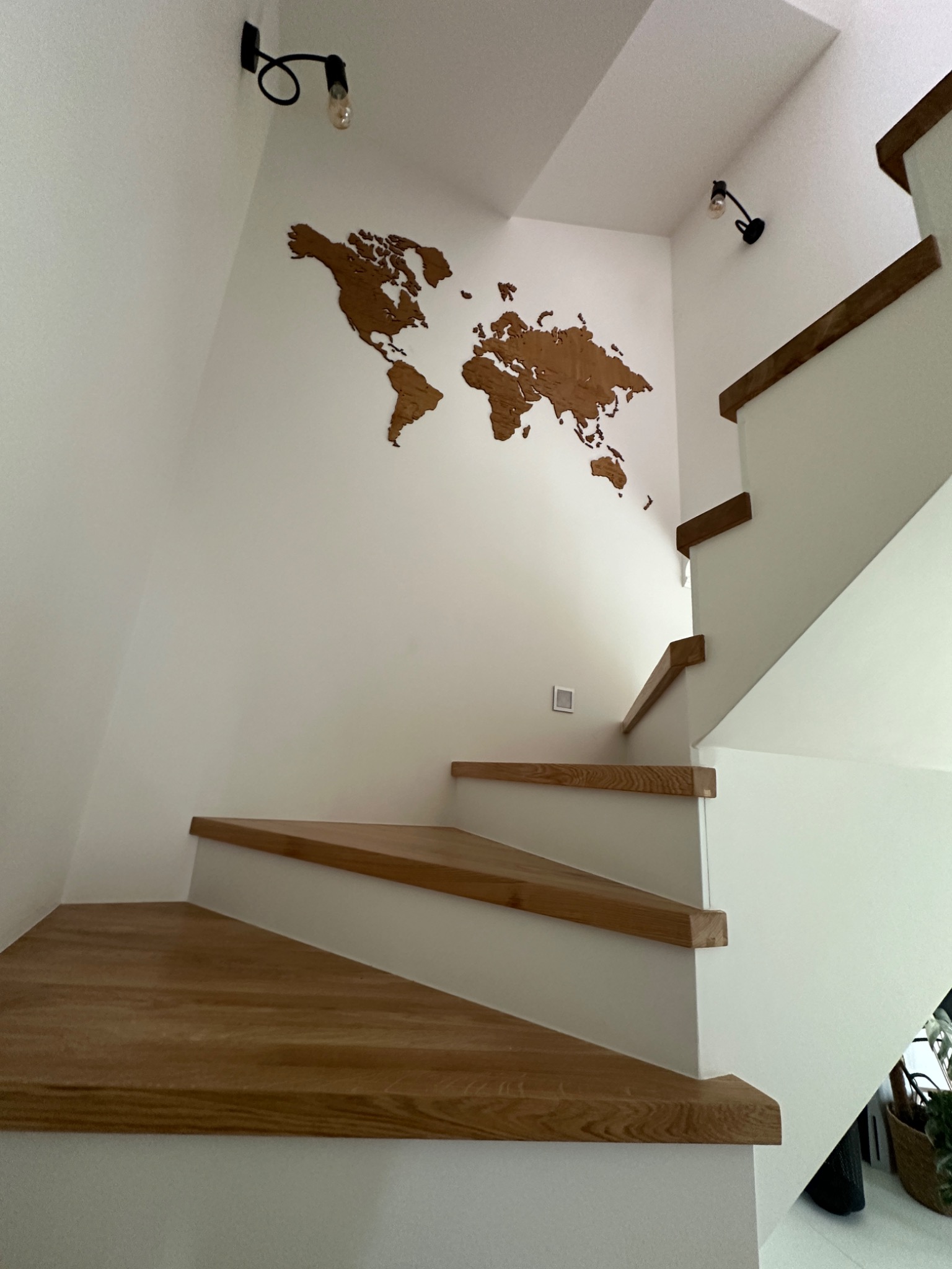 Nowoczesne schody drewniane z białymi podstopnicami, oświetlone kinkietami, z dekoracją ścienną w postaci drewnianej mapy świata.