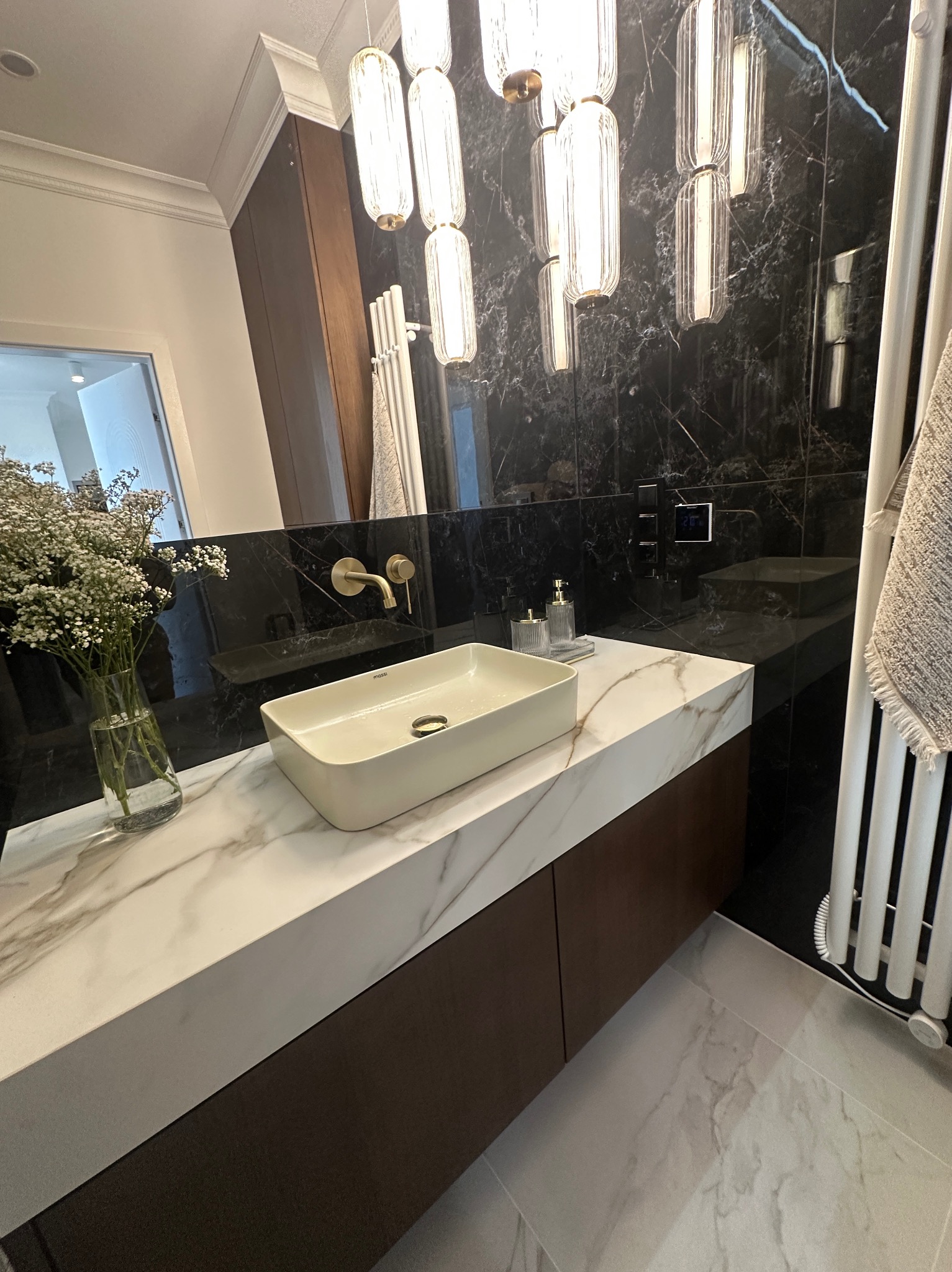 Nowoczesna łazienka z marmurową umywalką nablatową, czarnymi płytkami na ścianie, designerską lampą wiszącą i bukietem gipsówki w szklanym wazonie.