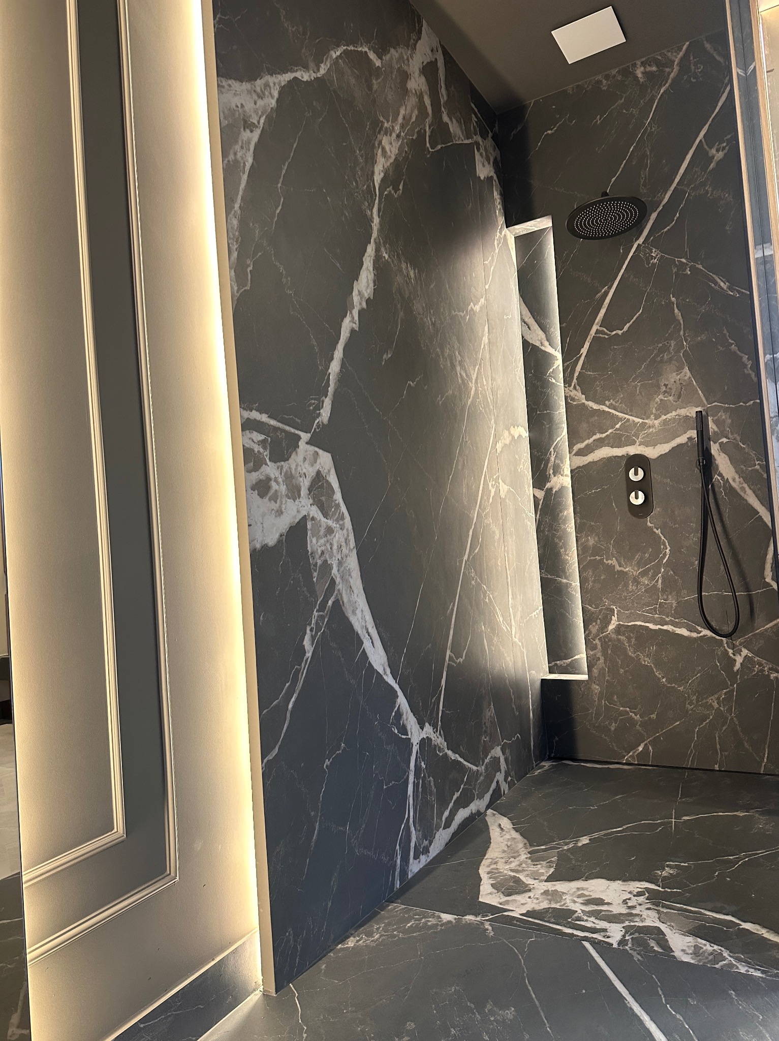 Nowoczesna kabina prysznicowa wykończona czarnym marmurem z białymi żyłami, oświetlona listwą LED, z czarną deszczownicą i baterią podtynkową.