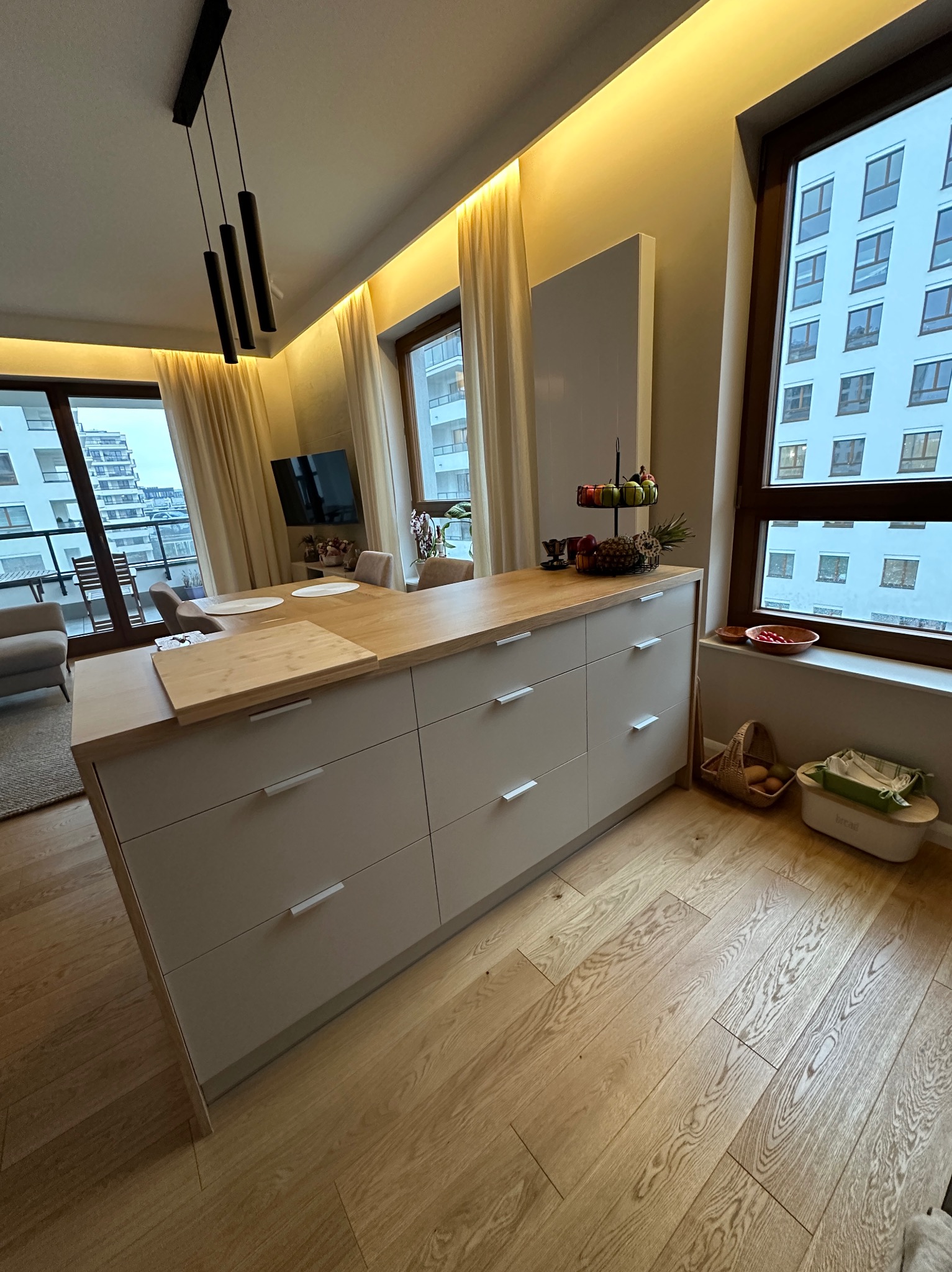 Jasna, nowoczesna kuchnia z wyspą, drewnianym blatem i podłogą, widok na balkon i zabudowę miejską za oknem.