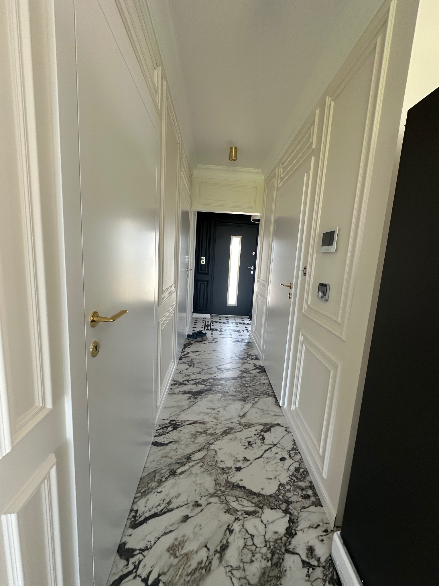 Korytarz z białymi drzwiami panelowymi, marmurową podłogą i czarnymi drzwiami wejściowymi w głębi.