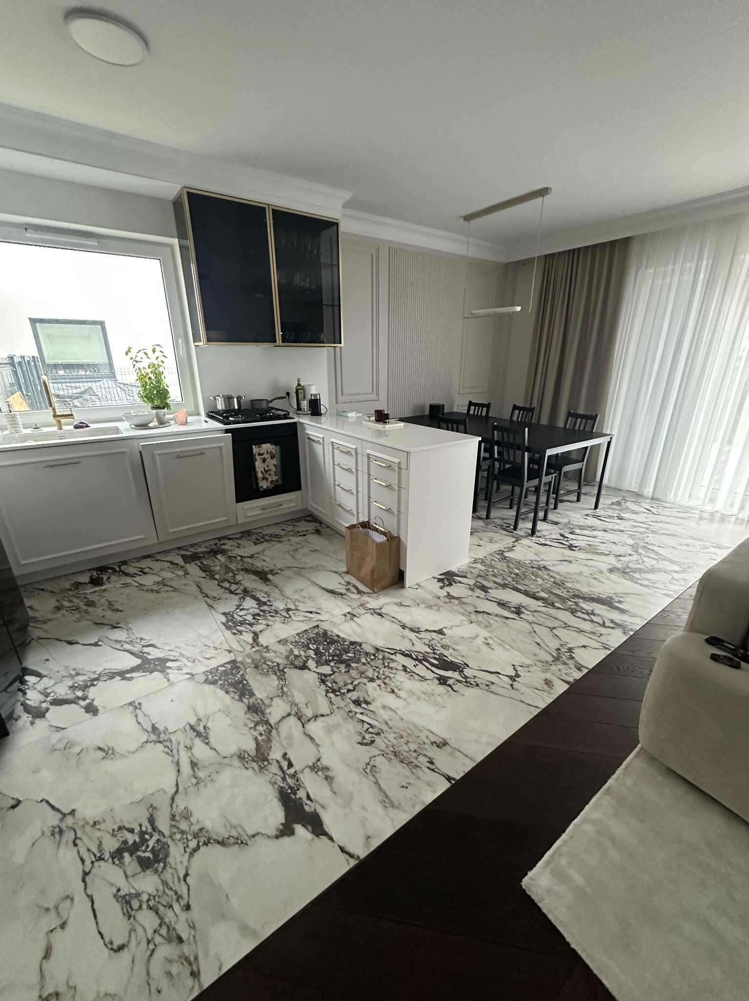 Nowoczesna kuchnia z marmurową podłogą, białymi szafkami i czarnym stołem jadalnym, doświetlona naturalnym światłem wpadającym przez okno.