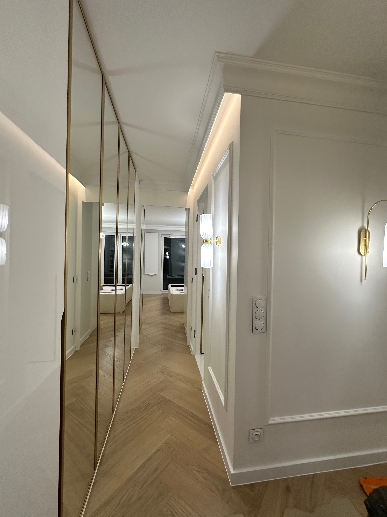 Nowoczesny korytarz z lustrzanymi szafami w złotych ramach, parkietem ułożonym w jodełkę i stylowym oświetleniem ściennym.