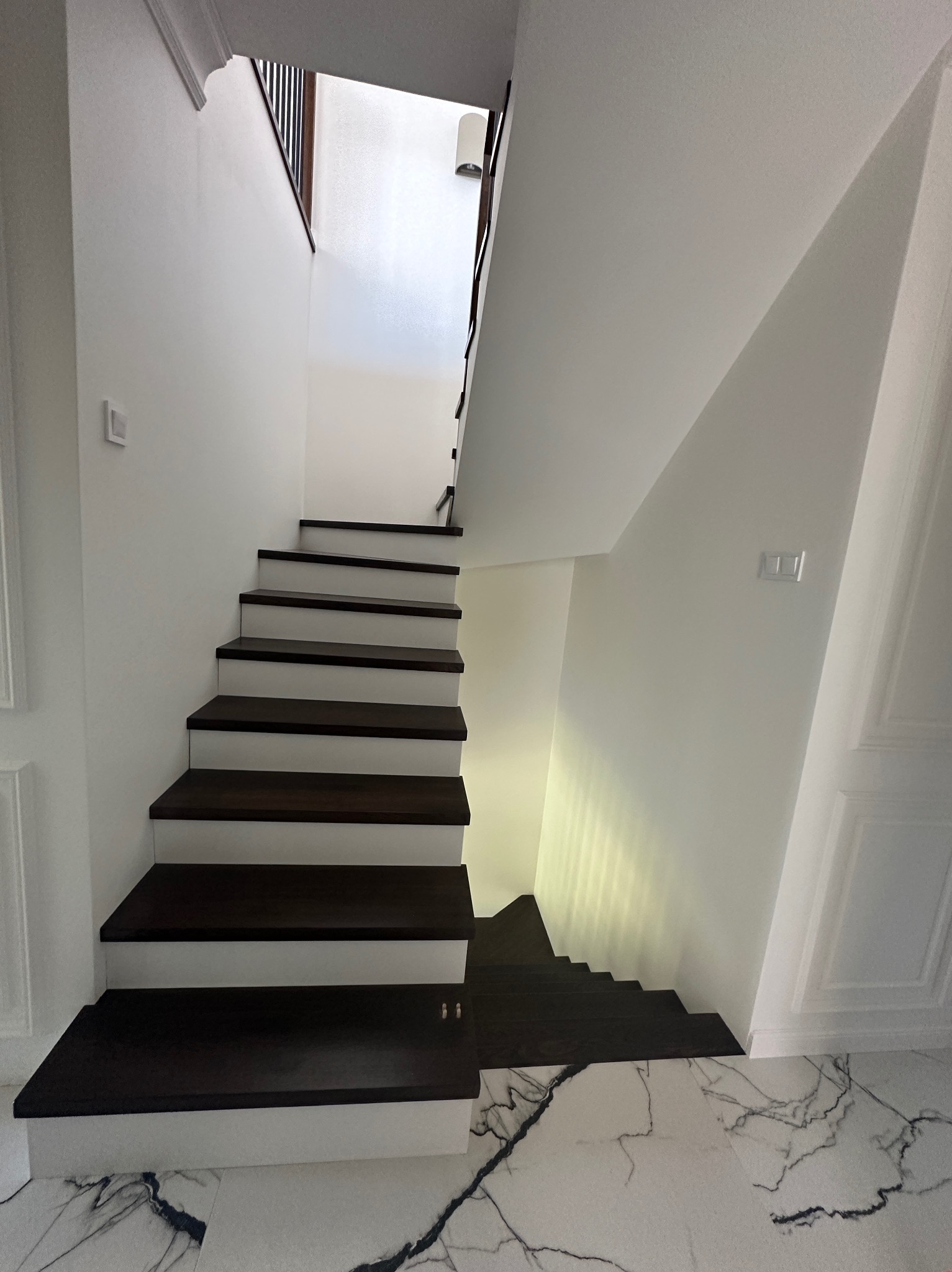 Nowoczesne schody wewnętrzne z ciemnego drewna na białych podstopnicach, marmurowa podłoga z czarnymi żyłami, białe ściany z dekoracyjnymi panelami.