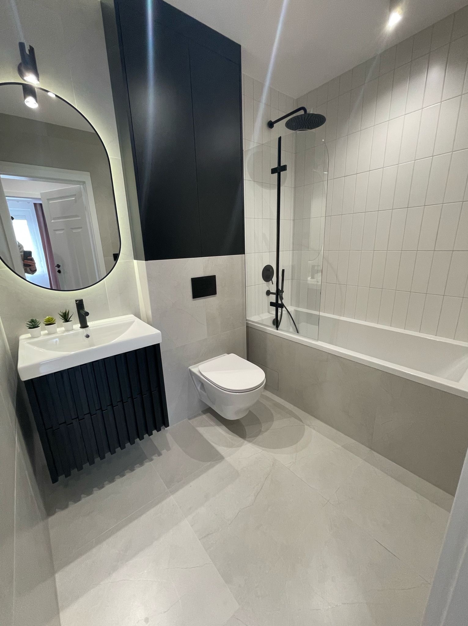 Nowoczesna łazienka z szarymi płytkami, czarną armaturą i szafką umywalkową z lamelami, owalne lustro z podświetleniem LED, minimalistyczny design.