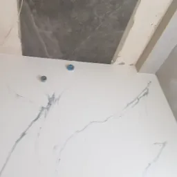 Biała ściana z szarymi żyłkami imitującymi marmur, widoczne dwa niebieskie i jeden srebrny element instalacji, surowy stan wykończenia ścian i sufitu w tle.