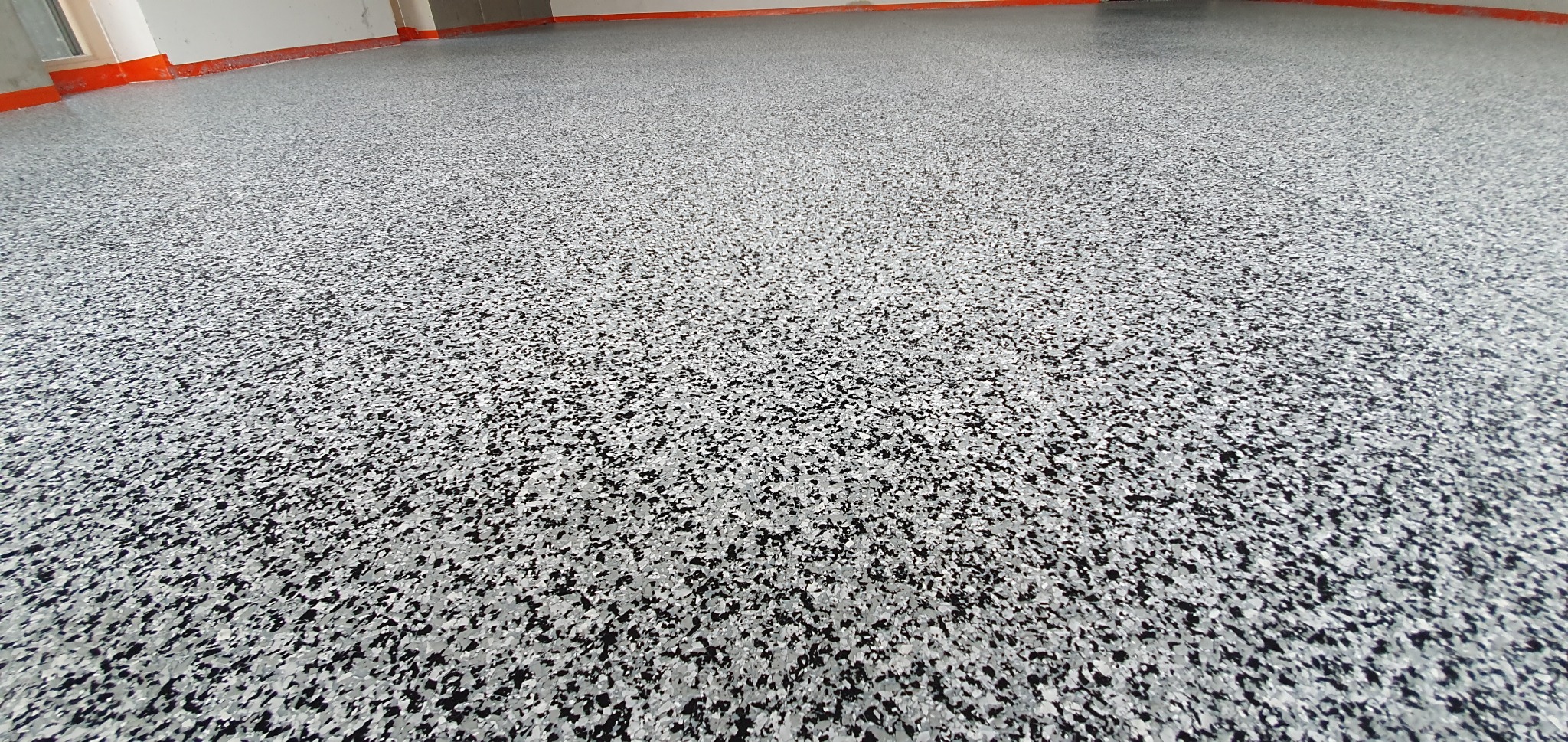 Szara posadzka żywiczna z czarnymi i białymi płatkami w garażu, widoczna pomarańczowa listwa przypodłogowa.