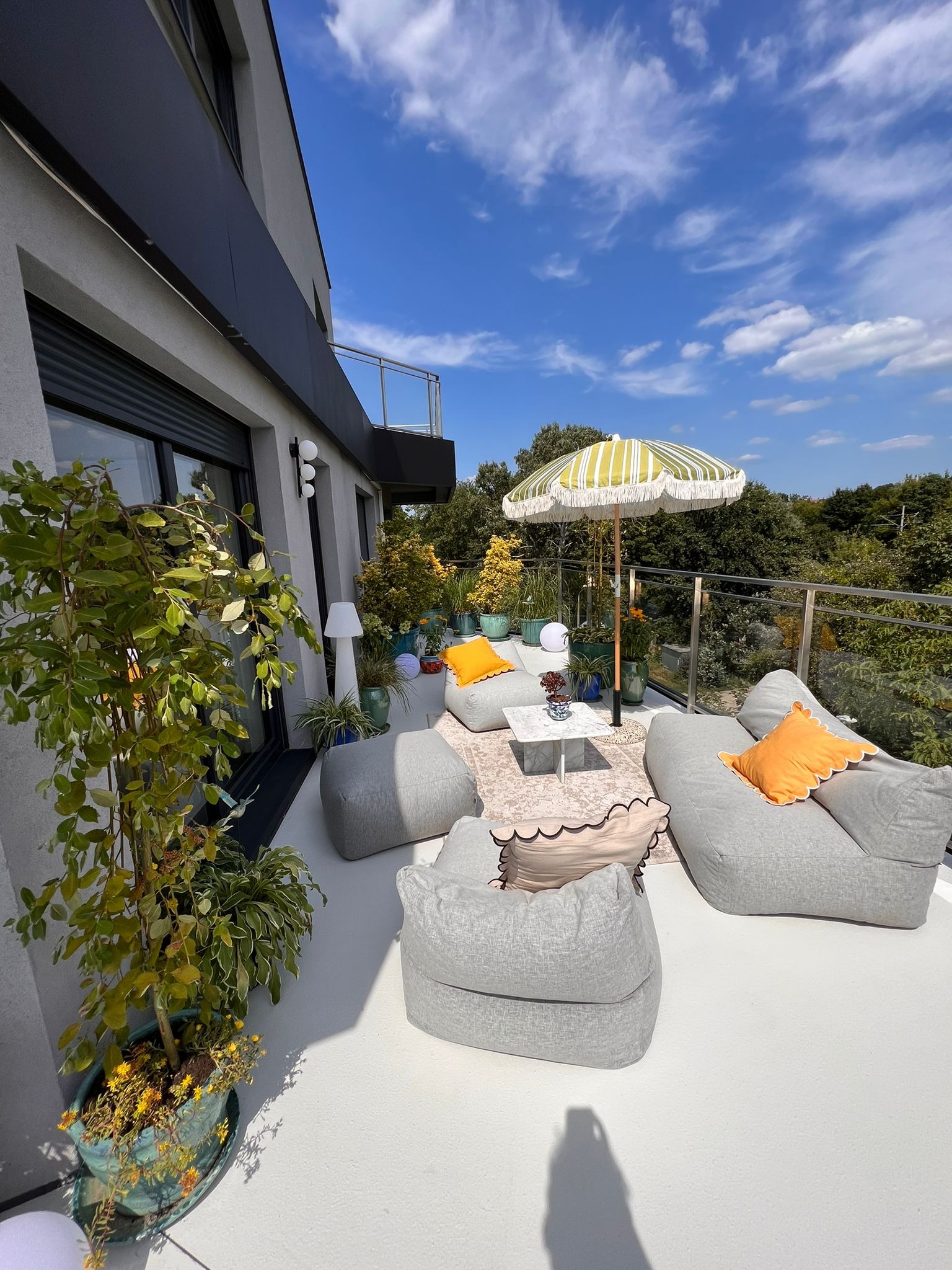 Jasnoszara, bezspoinowa powierzchnia balkonu z aranżacją wypoczynkową: pufy, marmurowy stolik, dywanik, parasol w paski i rośliny w ozdobnych donicach.