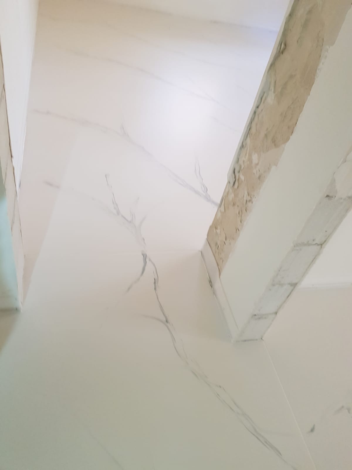 Biała posadzka z subtelnym marmurkowym wzorem, widoczny fragment surowego muru przy ścianie.