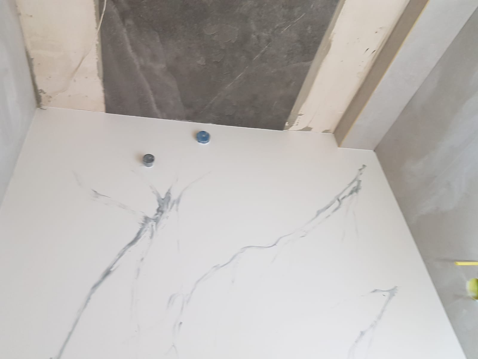 Biała ściana z szarymi żyłkami imitującymi marmur, widoczne dwa niebieskie i jeden srebrny element instalacji, surowy stan wykończenia ścian i sufitu w tle.