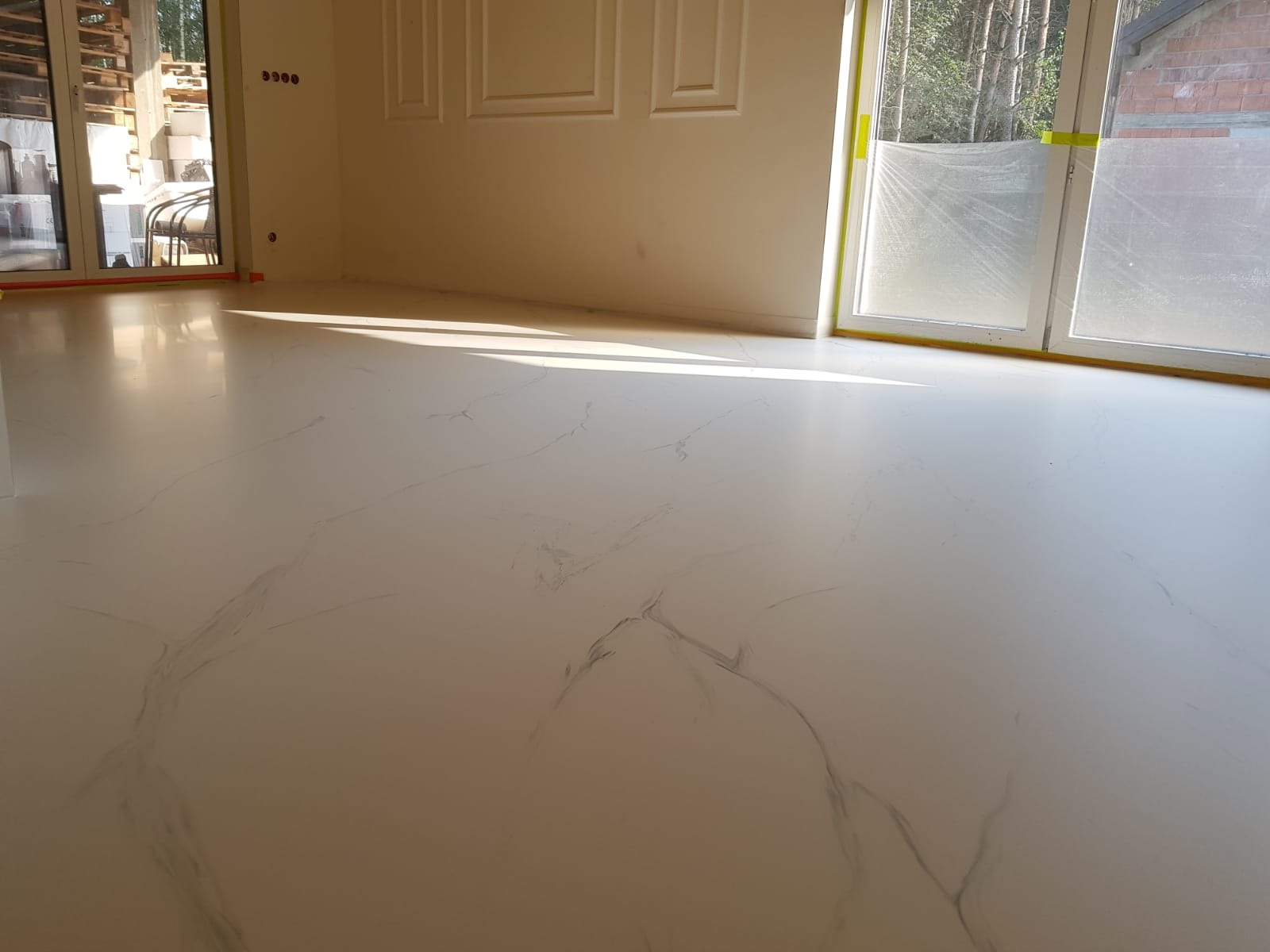 Błyszcząca, jasna podłoga z delikatnym marmurkowym wzorem w pustym pomieszczeniu z dużymi oknami zabezpieczonymi folią malarską.