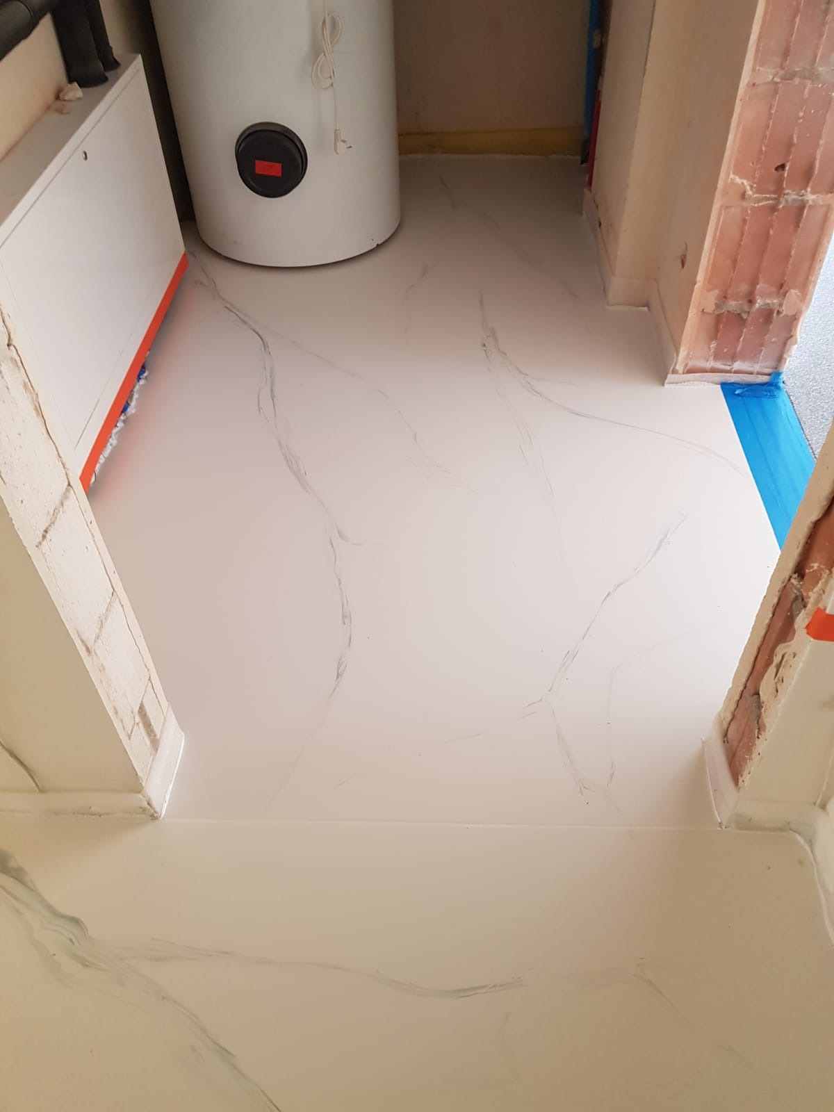 Jasna, marmurkowana podłoga w pomieszczeniu gospodarczym z bojlerem i instalacjami, zabezpieczona niebieską taśmą malarską przy progu.