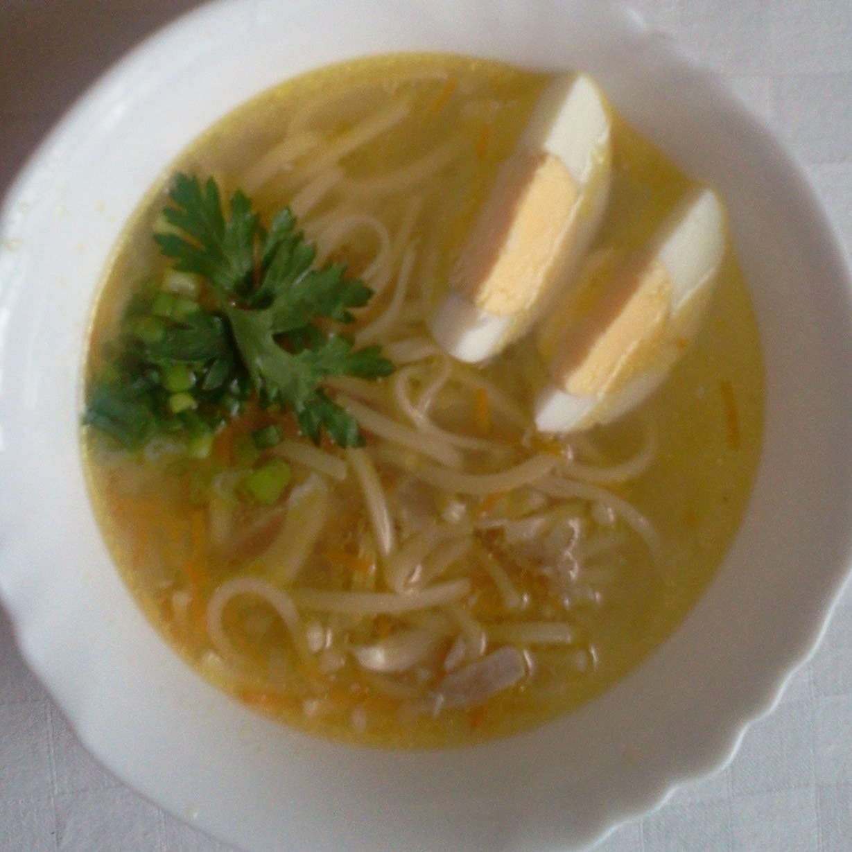 Apetyczna zupa rosołowa z makaronem, marchewką, zielonymi ziołami i dwoma połówkami jajka na twardo w białym talerzu.