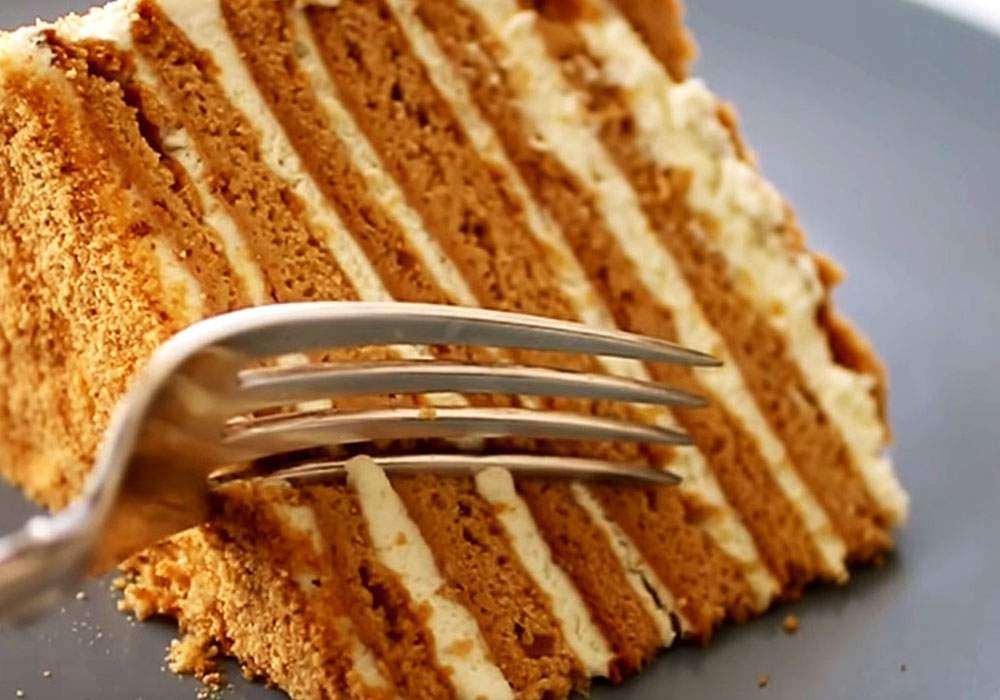 Kawałek wielowarstwowego tortu miodowego z kremem, nakłuty srebrnym widelcem na szarym talerzu.