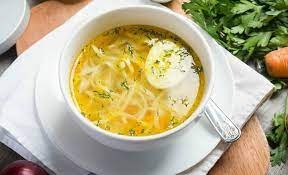 Rosół z makaronem i jajkiem w białym talerzu na drewnianej desce, obok świeża pietruszka, marchew i cebula.