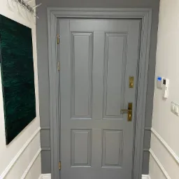 Szare, klasyczne drzwi wewnętrzne z mosiężnymi okuciami, zamontowane w ścianie z dekoracyjną sztukaterią i drewnianą podłogą.