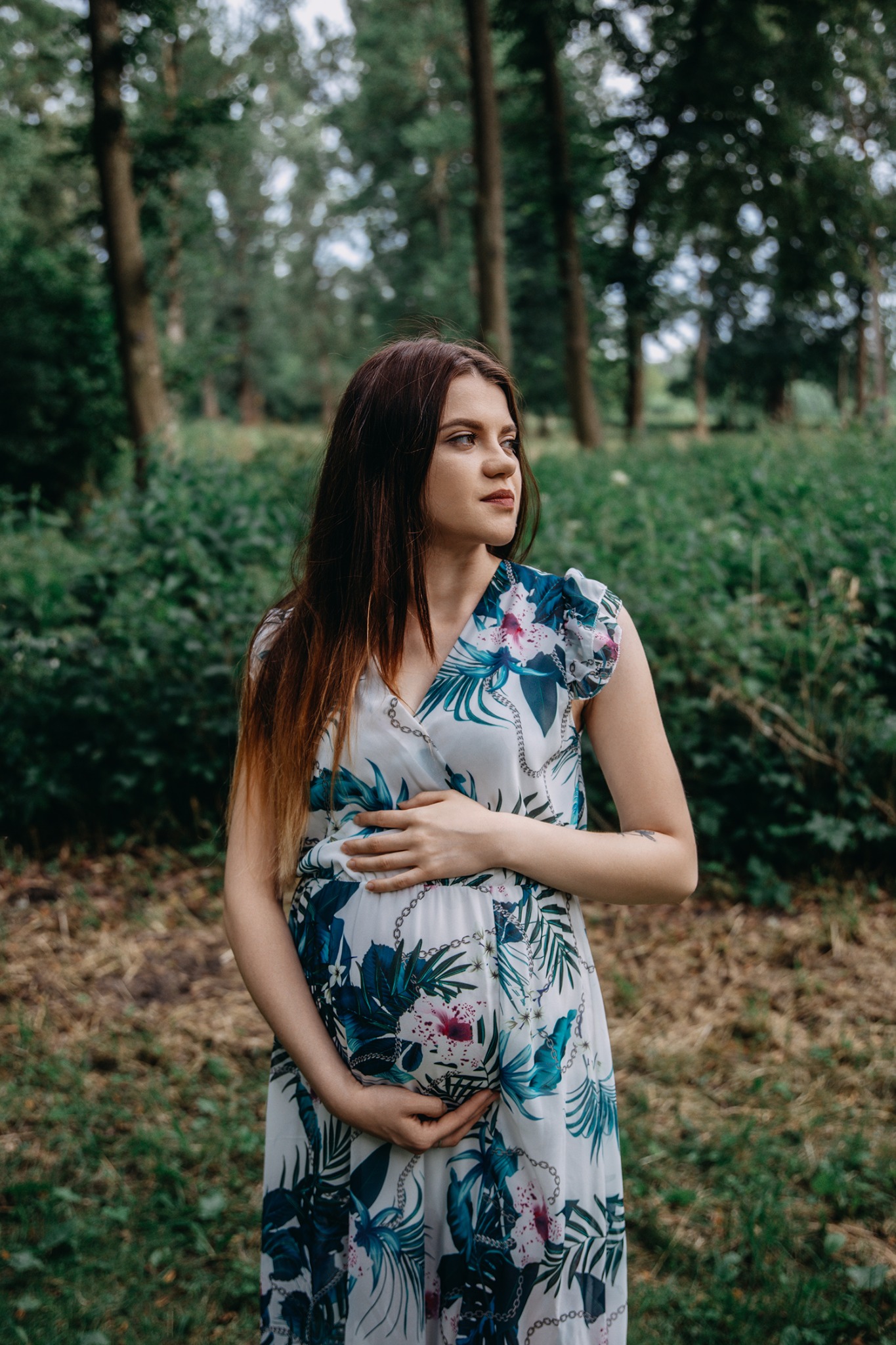 Kobieta w kwiecistej sukience ciążowej delikatnie obejmuje brzuch na tle leśnej zieleni.