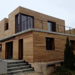 Nowoczesny dom z elewacją z drewnianych paneli, z balkonem na pierwszym piętrze, schodami wejściowymi i osobą siedzącą na krawędzi dachu sąsiedniego budynku.