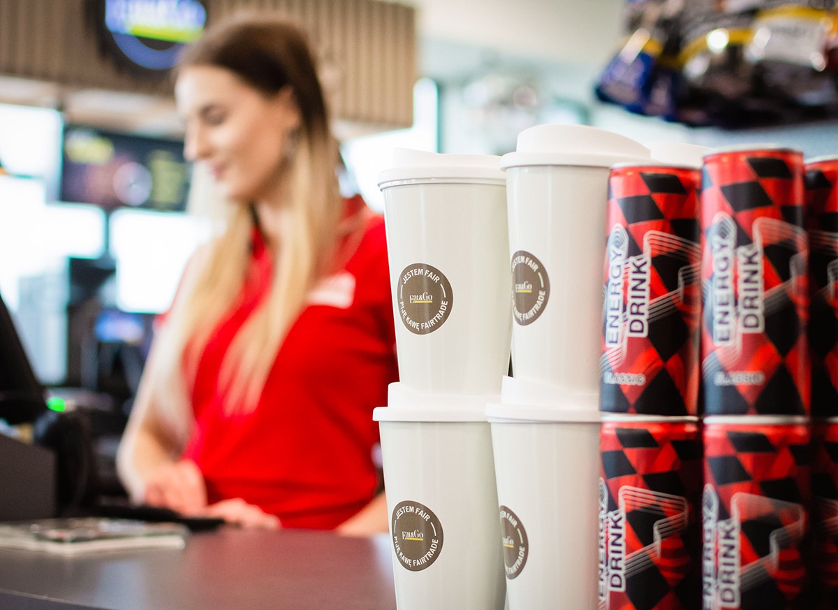 Stos kubków z logo 'Jestem Fair' obok puszek napojów energetycznych, w tle rozmyta postać kobiety w czerwonej koszulce przy kasie, wnętrze punktu sprzedaży.