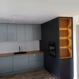 Nowoczesna kuchnia z frontami w kolorze gołębim, drewnianym blatem i czarną armaturą, uzupełniona o ciemnoszarą zabudowę z piekarnikiem i podświetlanymi półkami z jasnego drewna.