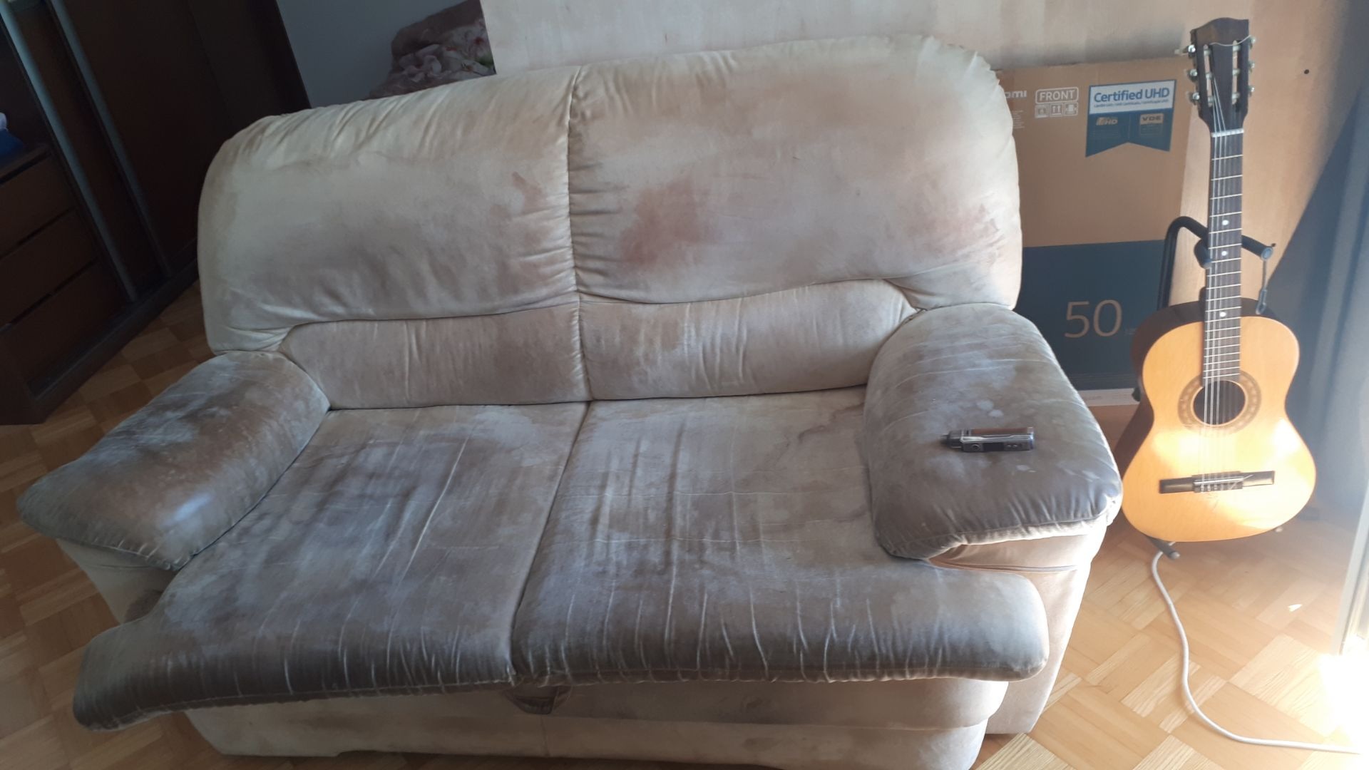 Używana, dwuosobowa sofa z plamami i śladami użytkowania, obok gitara klasyczna i karton po telewizorze.