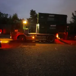 Ciężarówka z dwoma kontenerami na platformie, widoczna w słabym oświetleniu wieczornym, z włączonymi światłami pozycyjnymi.